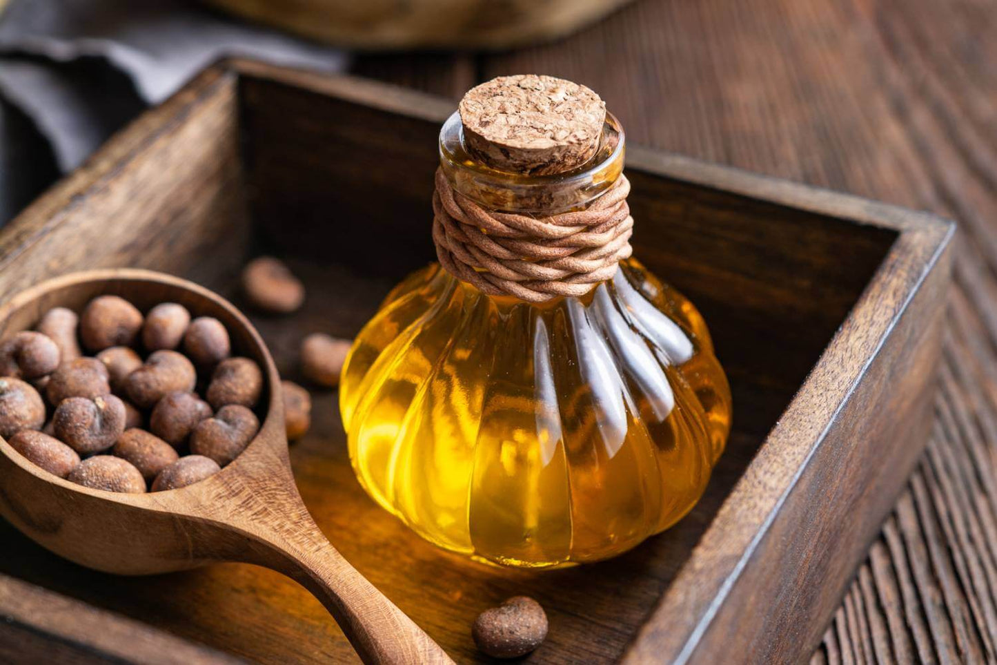 Baobab Oil in a round glass bottle with a cork and twine around the necl. There is a wooden spoon filled with Baobab seeds both in a wooden crate on a wooden surface 