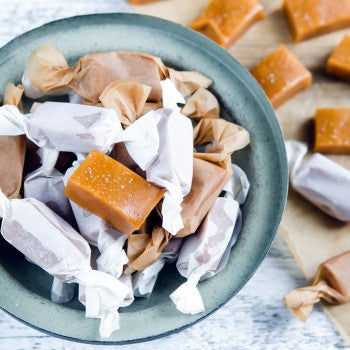 one unwrapped square butterscotch sweet, several butterscotch sweets wrapped in whit paper in a sky blue bowl, unwrapped butterscotch sweets on the right side of the bowl with two wrapped butterscotch sweets all on a white table cloth