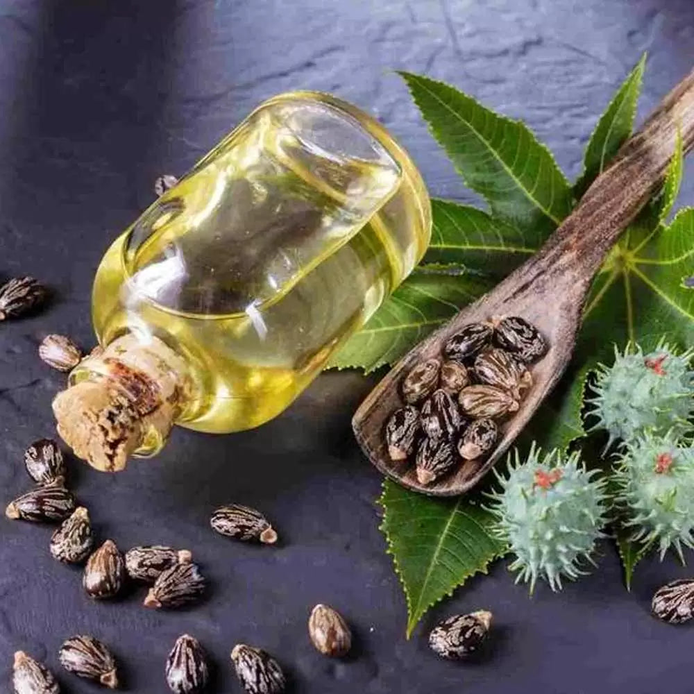 Castor Oil in a transparent glass bottle with cork lid on a black surface with a wooden spoon of castor seeds and leaves