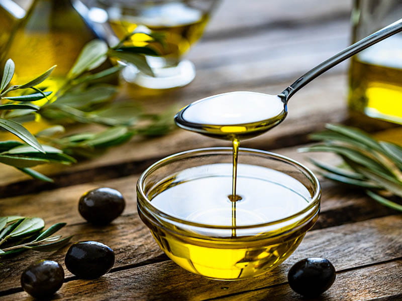 spoon full of olive oil over a glass bowl of olive oil on a wooden surface with fresh black olives and olive leaves