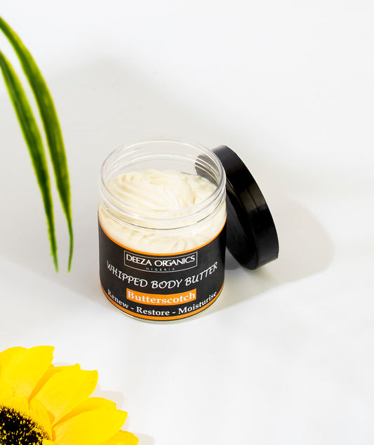 Whipped Butterscotch body butter in a open transparent jar with its black lid leaning on the jar on a with background with sunflower and leaf props 
