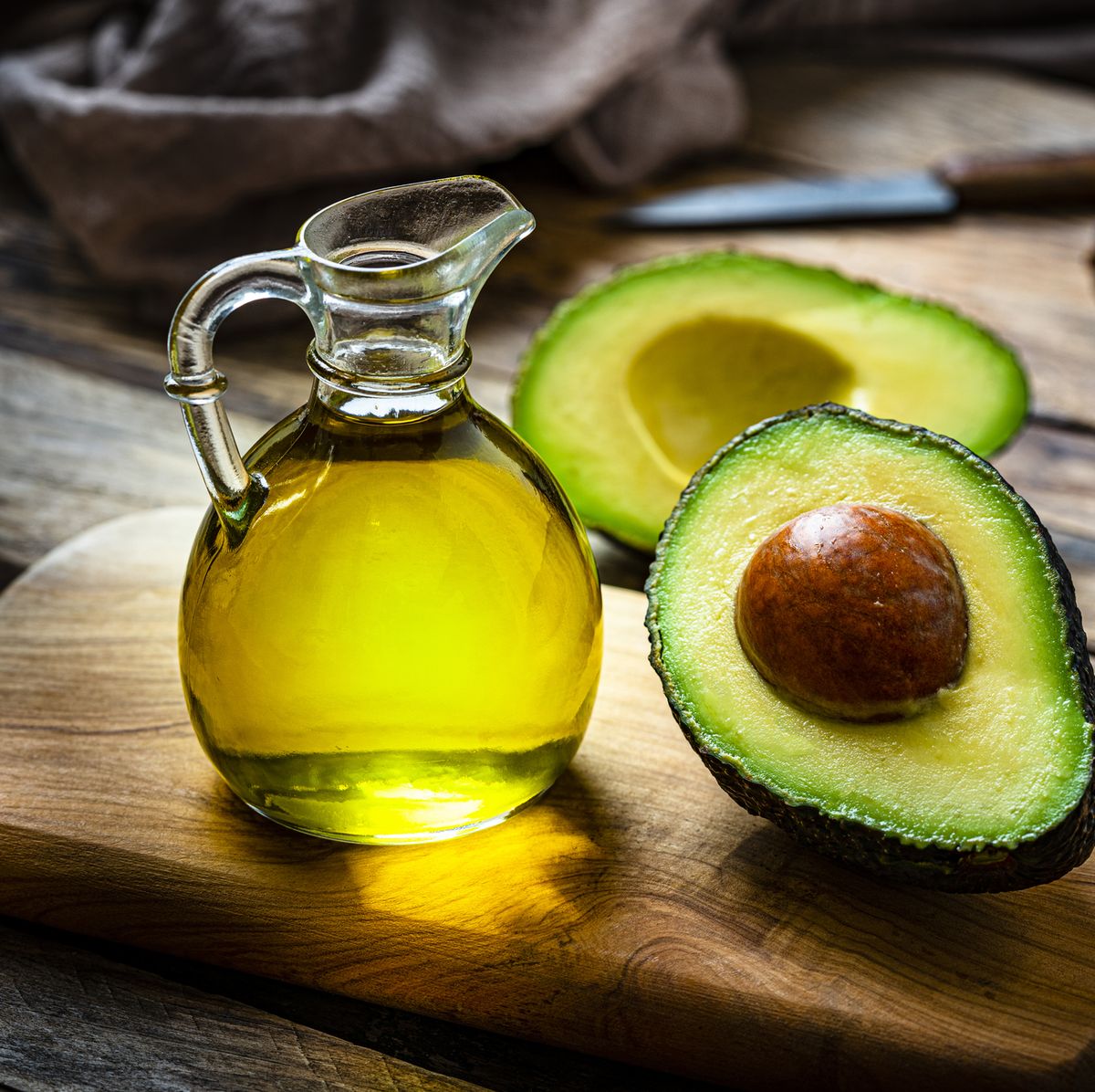 Avocado oil in a transparent glass jug with opened avocado fruit on a wooden chopping board. Fruit knife inn the corner to the rigt