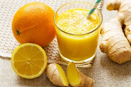 Orange Juice in a transparent glass cup with a white and blue straw in it. bHalf cit and full orange with split  ginger root on a table 