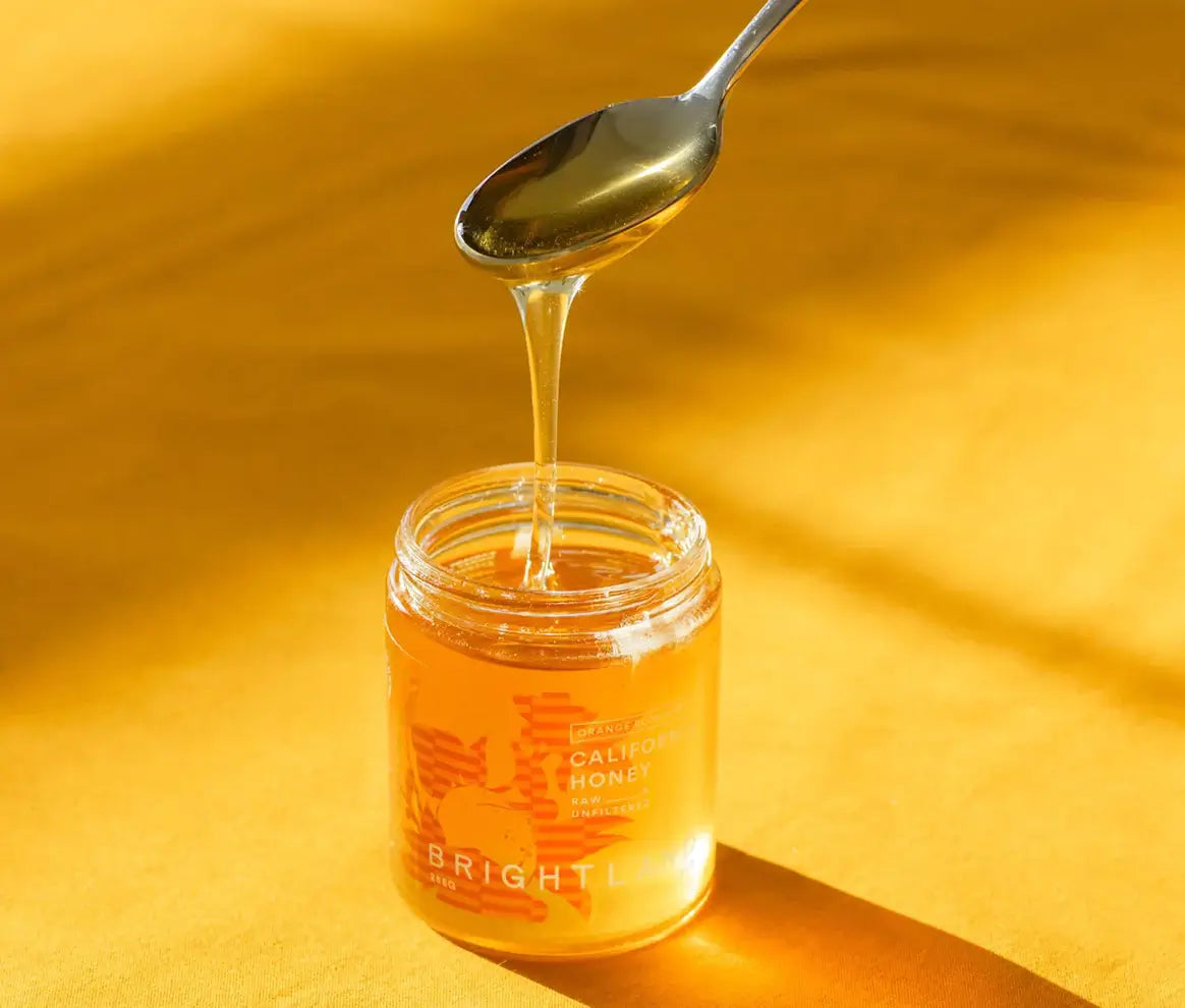 A silver spoon pouring honey into a full jar of honey . Honey jar is transparent and on a yellow surface