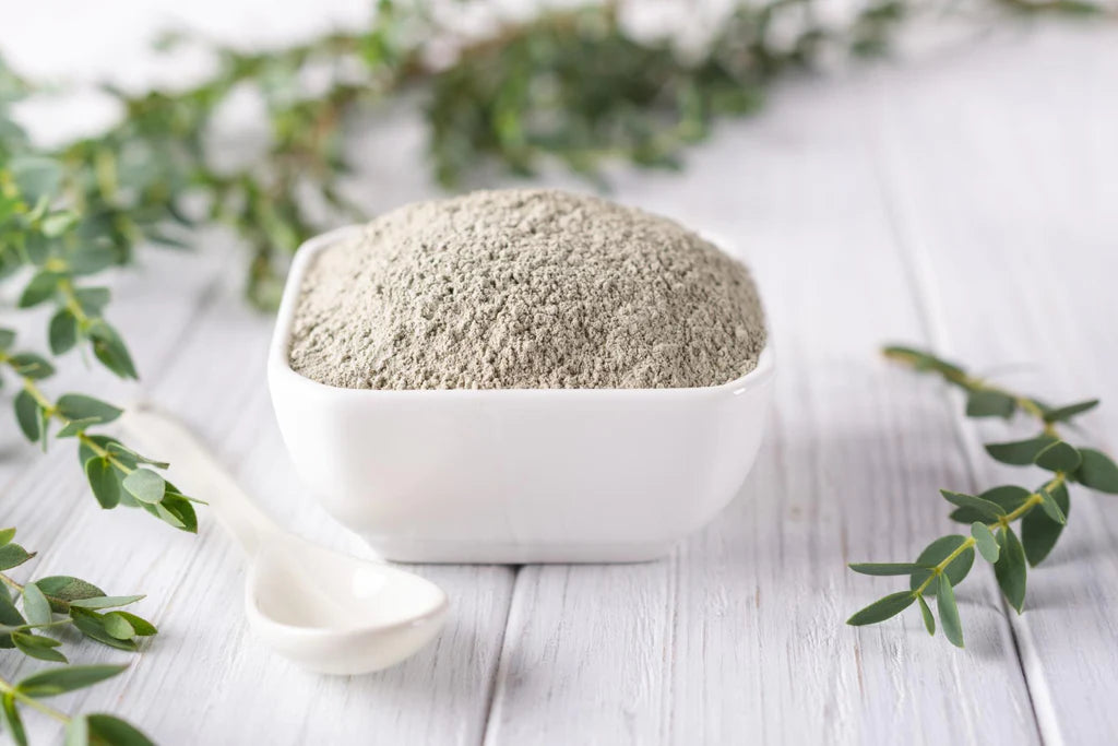 Bentonite clay in a white square bowl with a white spoon by it's side both on a white wooden surface with leaves 